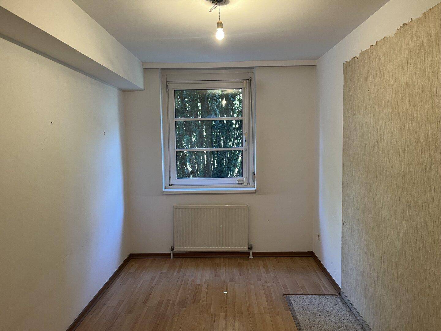 Leeres Zimmer mit teilweise gestreifter Tapete, Holzfußboden und einem Fenster mit Rollladen.