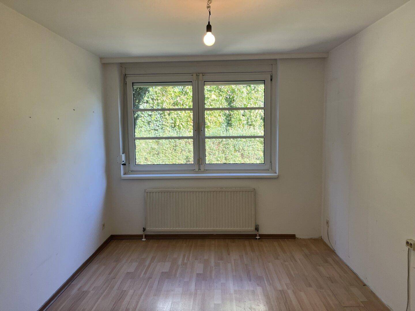 Leeres Zimmer mit Holzfußboden, weiß gestrichenen Wänden und einem Fenster mit Rollladen.