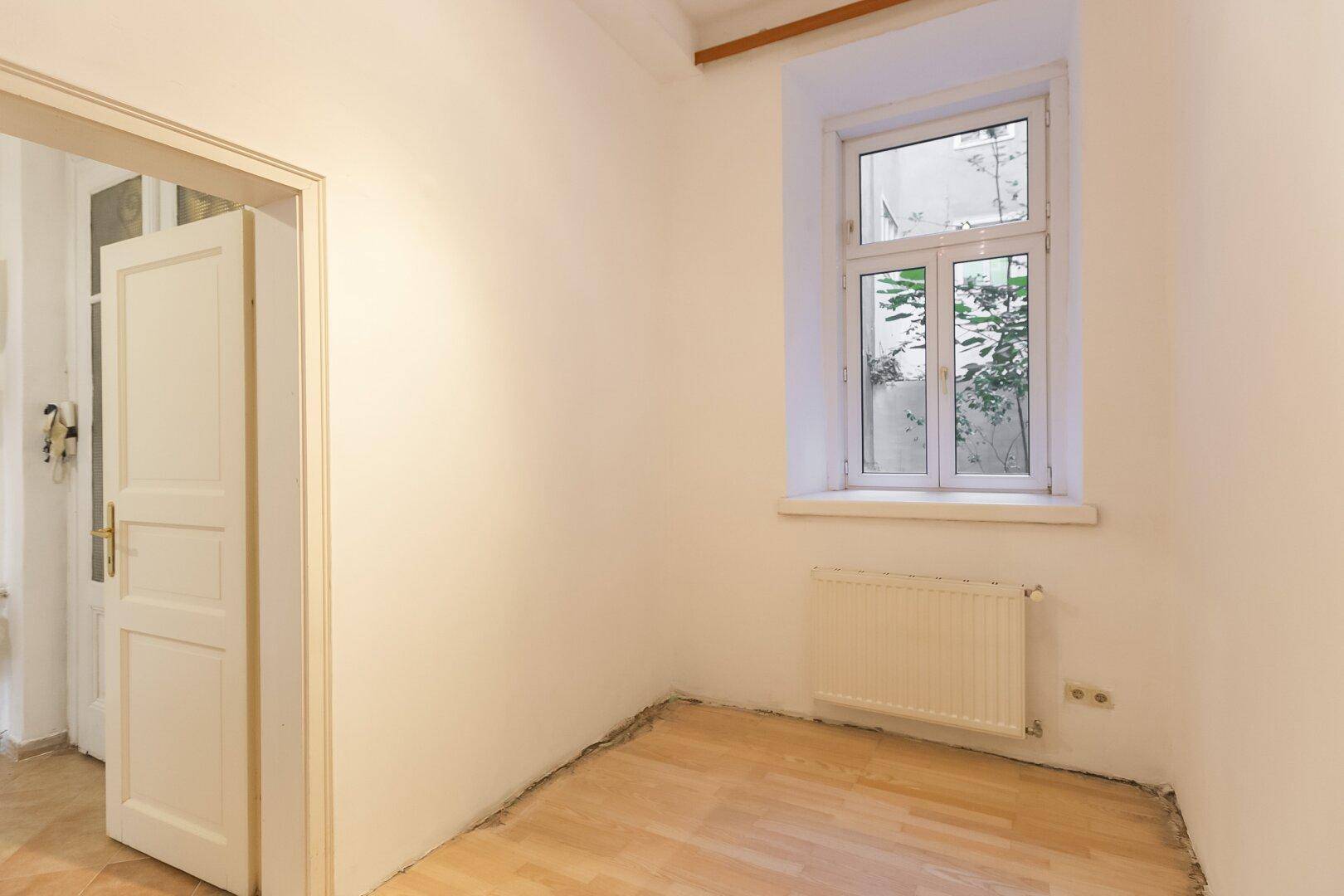 Kleines, helles Zimmer mit Holzboden und Blick auf einen ruhigen Innenhof.