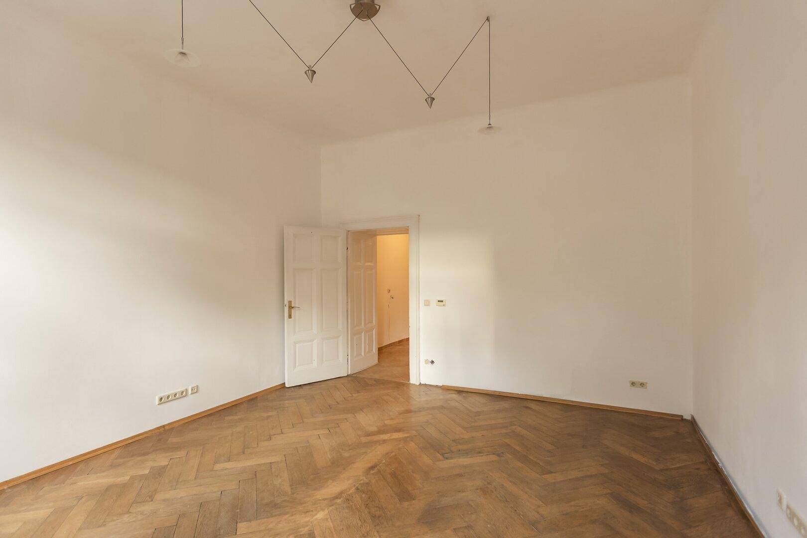 Geräumiges Zimmer mit klassischem Fischgrätparkett und einer Doppeltür zu einem weiteren Raum.
