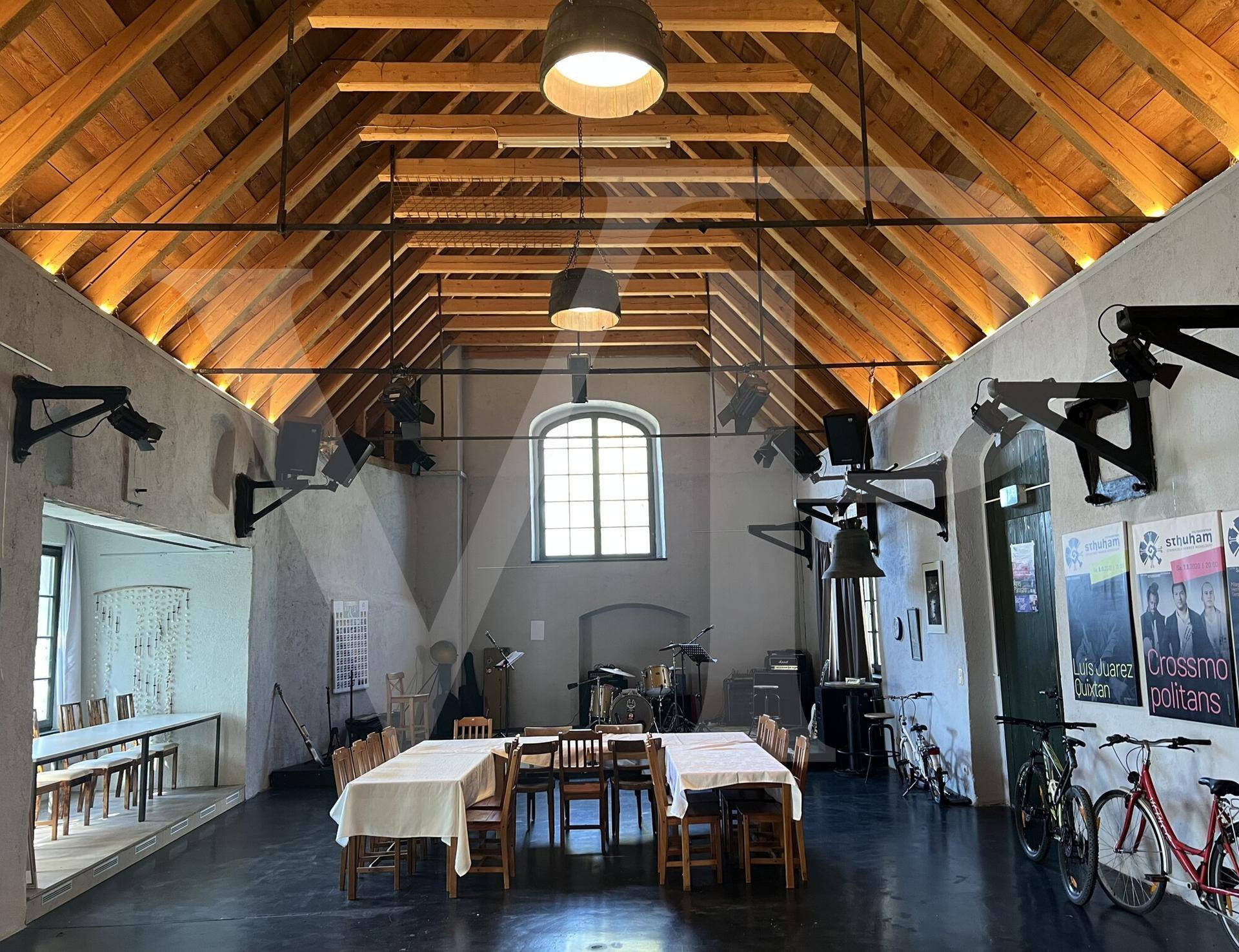 Großer Saal mit hoher Holzdecke, Bühne und Tischen, ideal für Veranstaltungen.