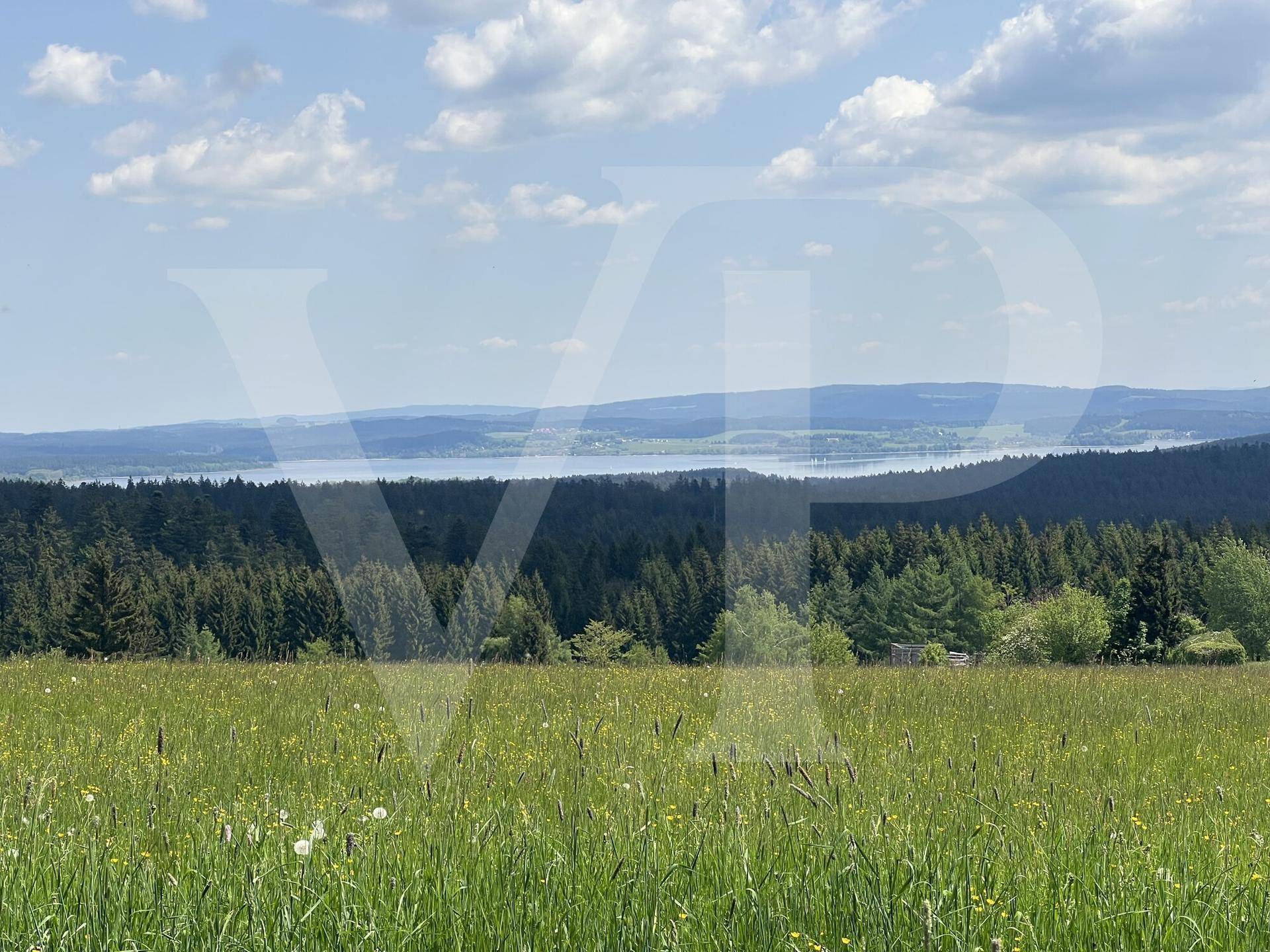 Weitläufige Landschaft mit grünen Wiesen, Wäldern und einem See im Hintergrund unter blauem Himmel.