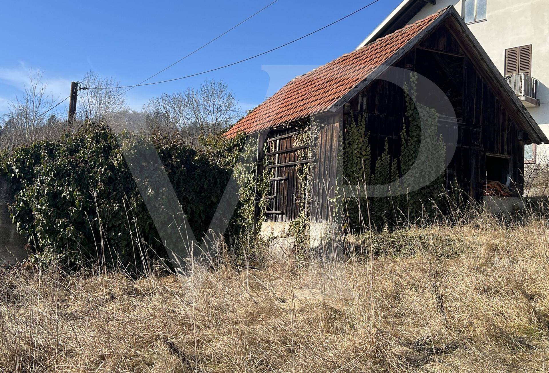 Kleines, verfallenes Nebengebäude mit Holzwänden und Ziegeldach, umgeben von Vegetation.
