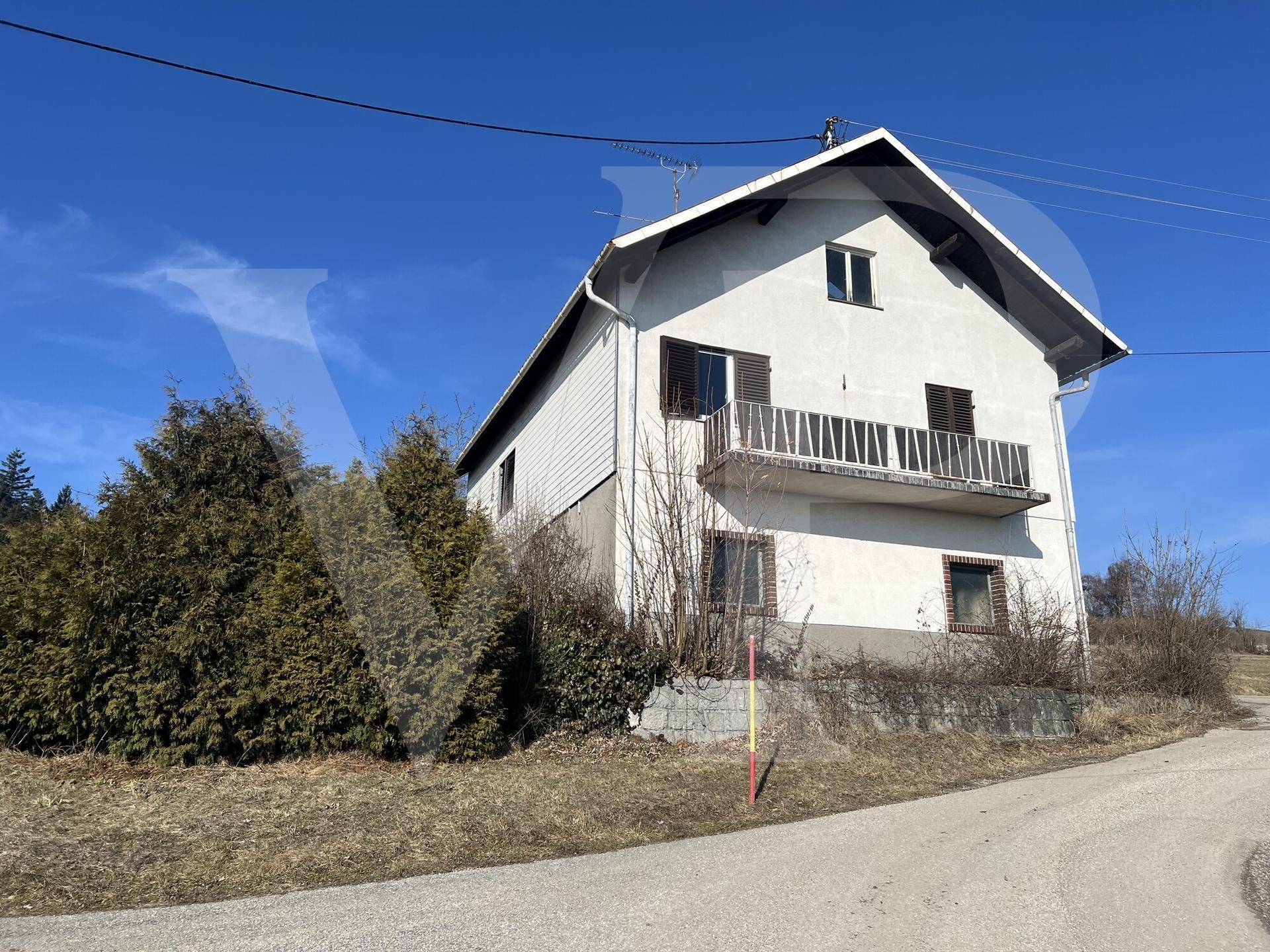 Einfamilienhaus mit Balkon und grüner Hecke im Vordergrund, gelegen in einer sonnigen Landschaft.