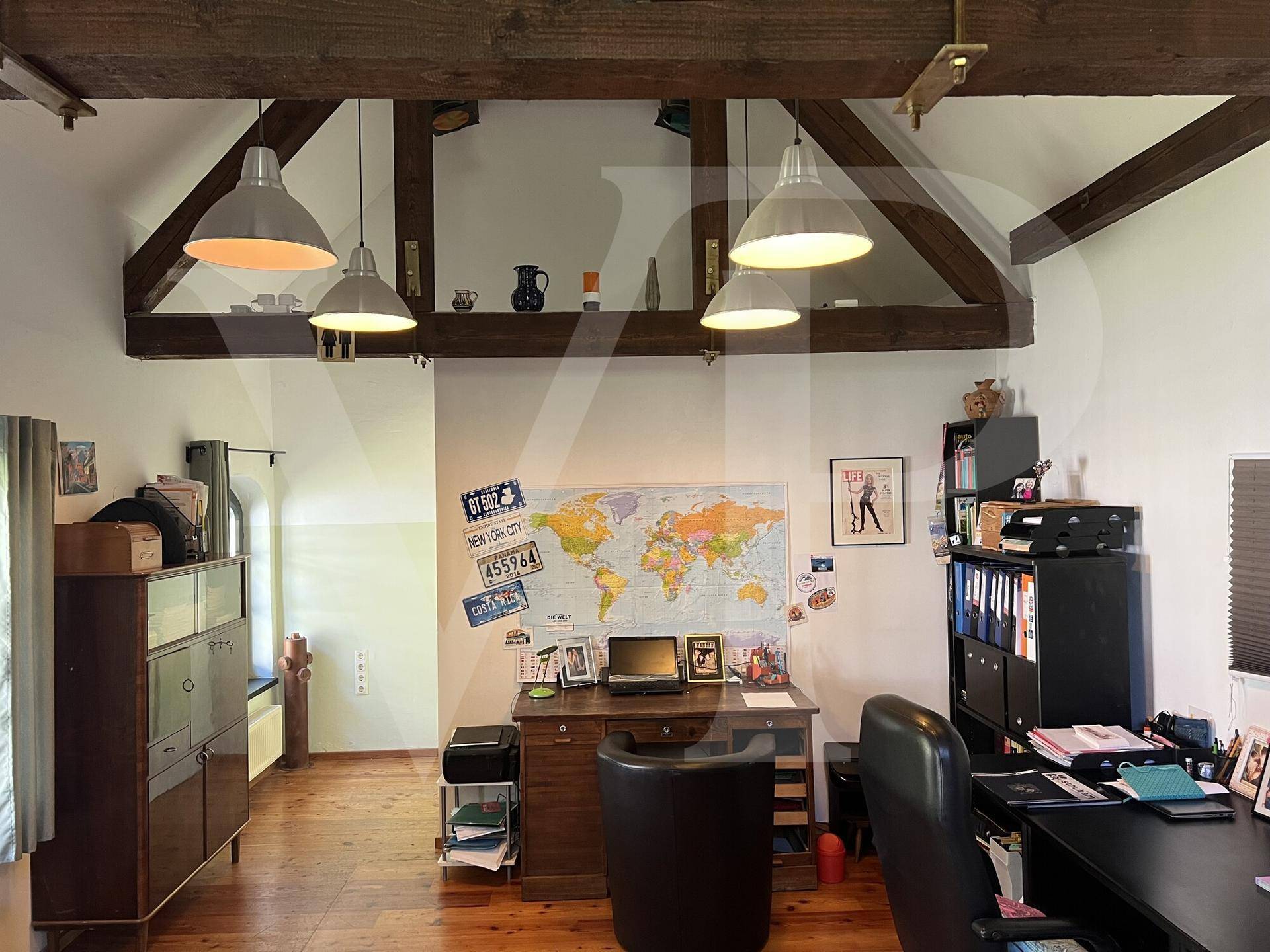 Helles Büro mit Schreibtisch, Regalen und einer Weltkarte an der Wand, unter Holzbalken.