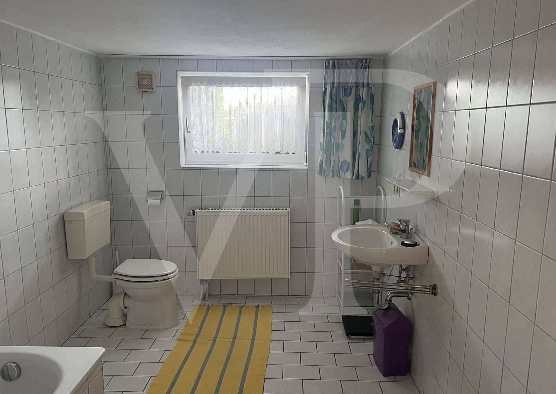 Badezimmer mit weißen Fliesen, Fenster, Toilette und Waschbecken im Stil der 80er Jahre.