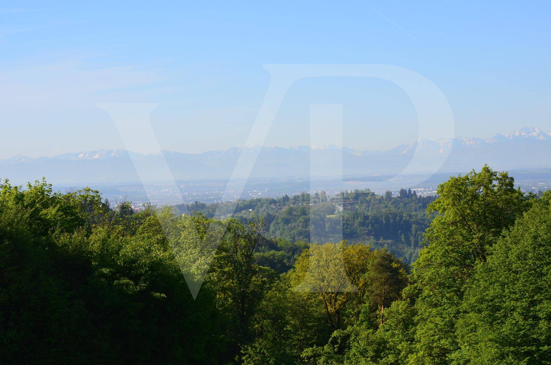 Blick über eine hügelige, bewaldete Landschaft mit einer Siedlung in der Ferne und Bergen am Horizont.