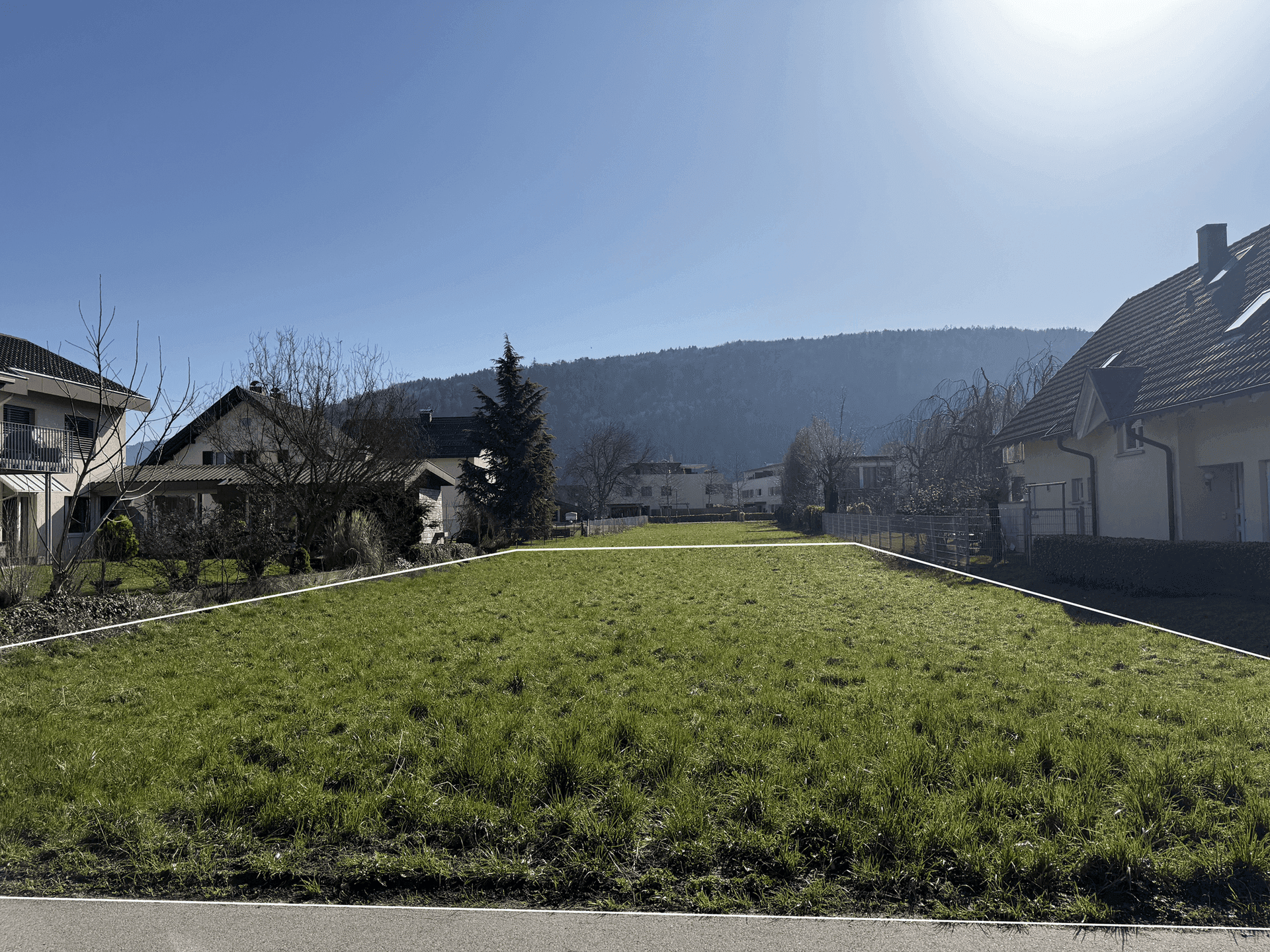 Freies Baugrundstück mit grüner Grasfläche, umgeben von bestehenden Wohnhäusern und Hügeln.