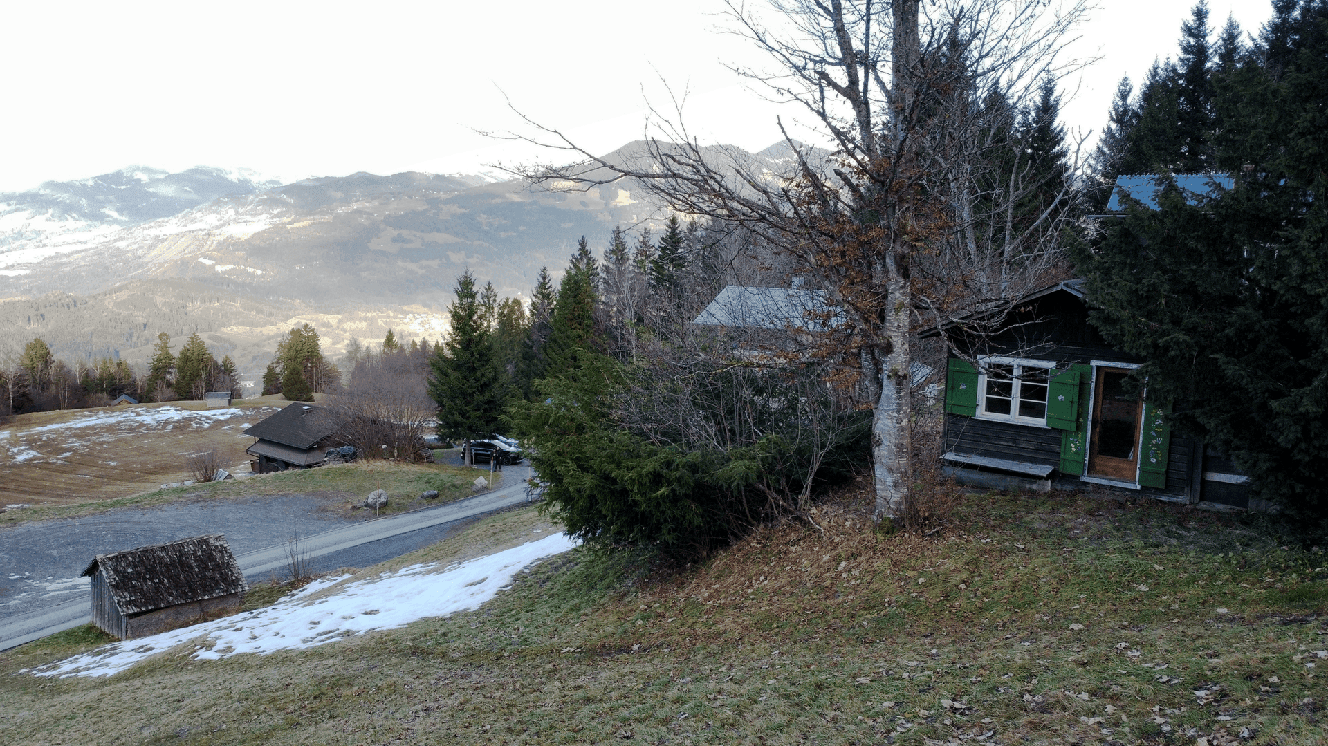 Blick auf die rustikale Holzhütte und die umliegende Berglandschaft mit Straße und weiteren Gebäuden.