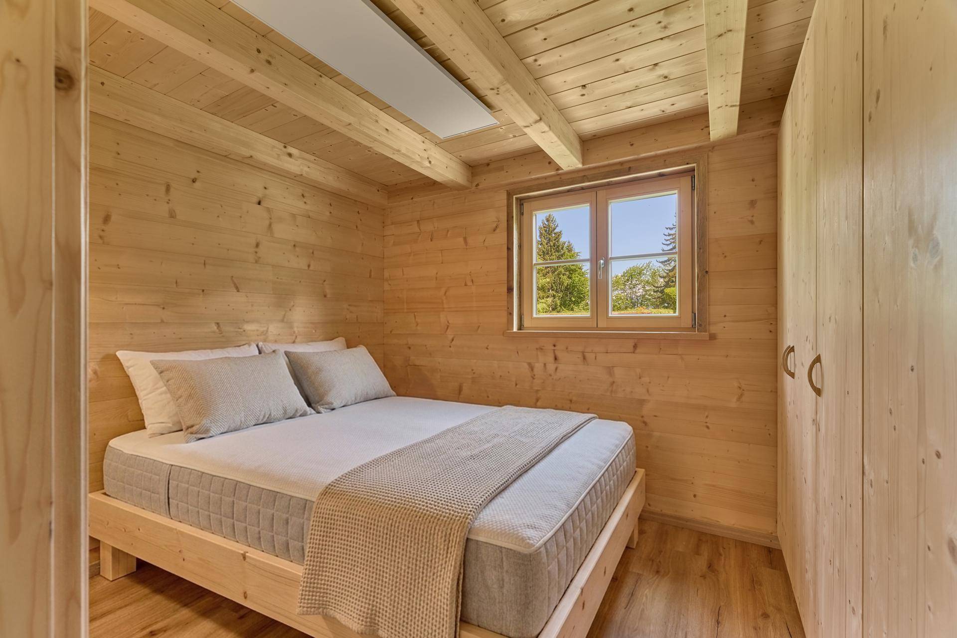 Helles Schlafzimmer in einem Holzchalet mit Holzwänden, Doppelbett und Fensterblick ins Grüne.