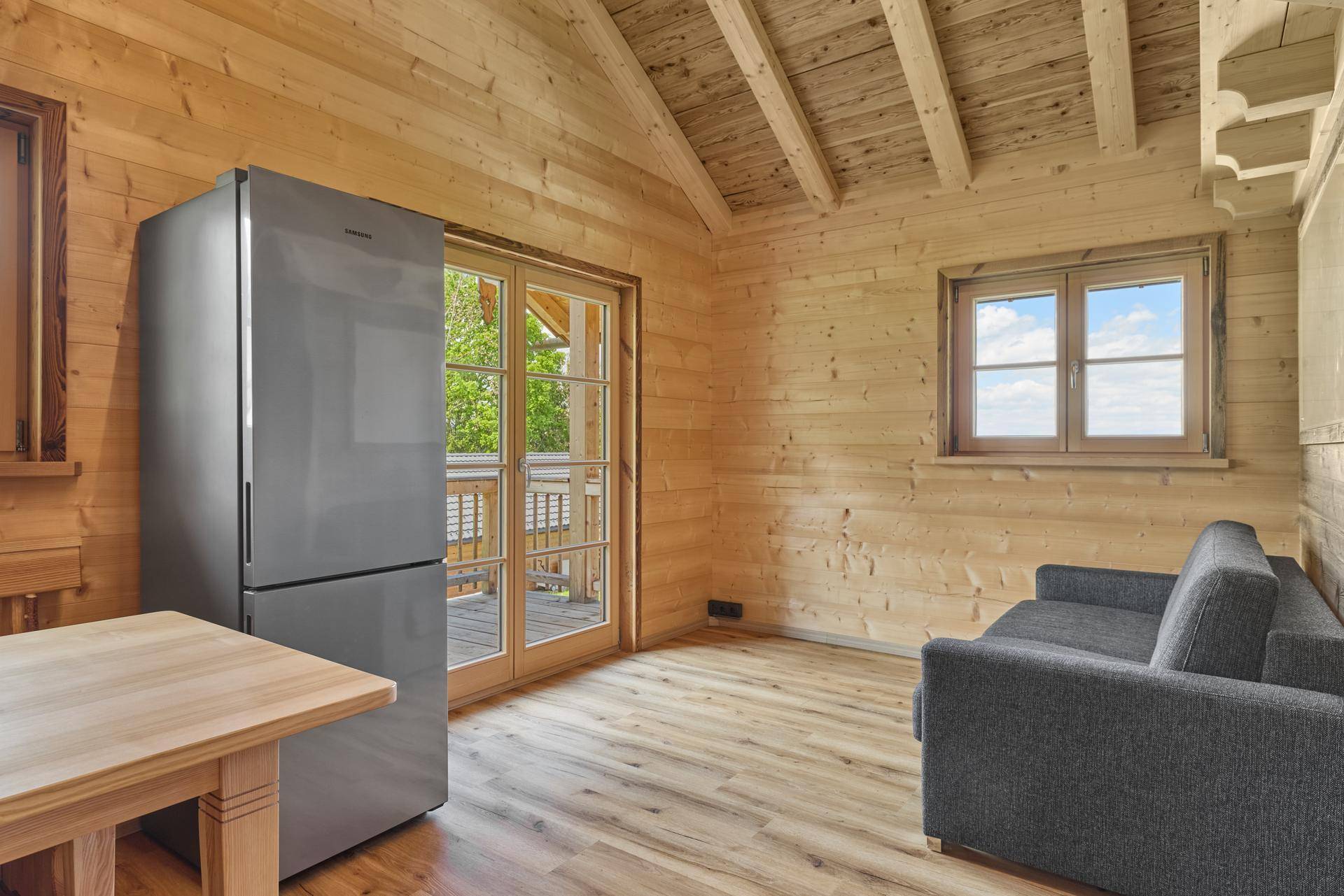 Gemütlicher Wohnbereich in einem Holzchalet mit Sofa, Kühlschrank und Balkonzugang.