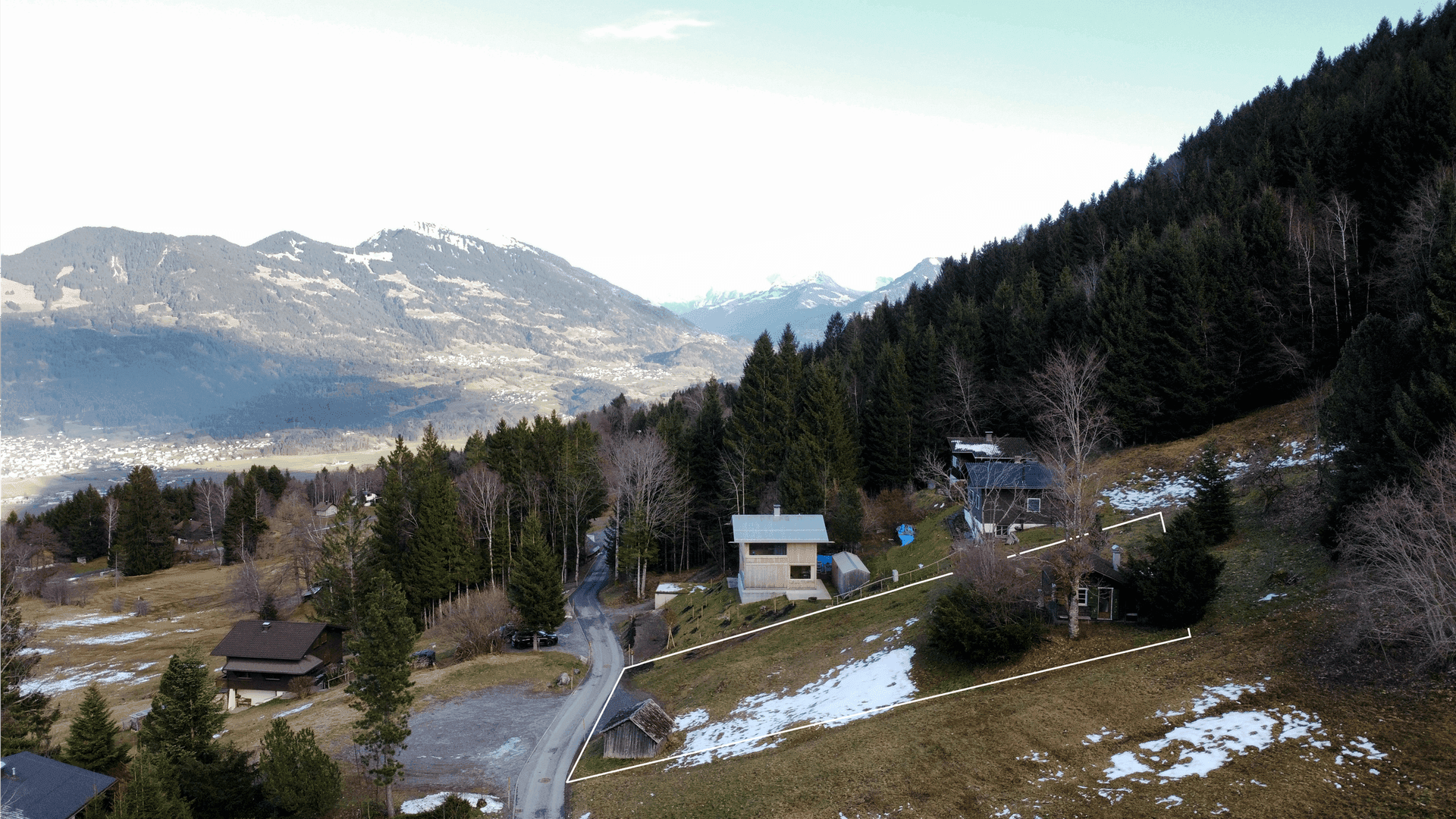 Weitläufige Luftaufnahme einer Berglandschaft mit verstreuten Häusern und bewaldeten Hängen.