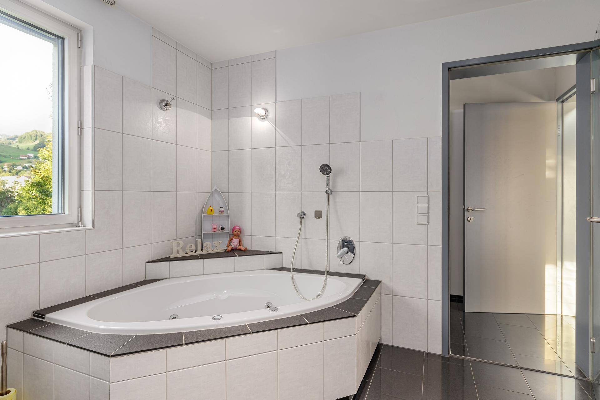 Helles Badezimmer mit Eckbadewanne, Fenster und modernen weißen Fliesen an den Wänden.