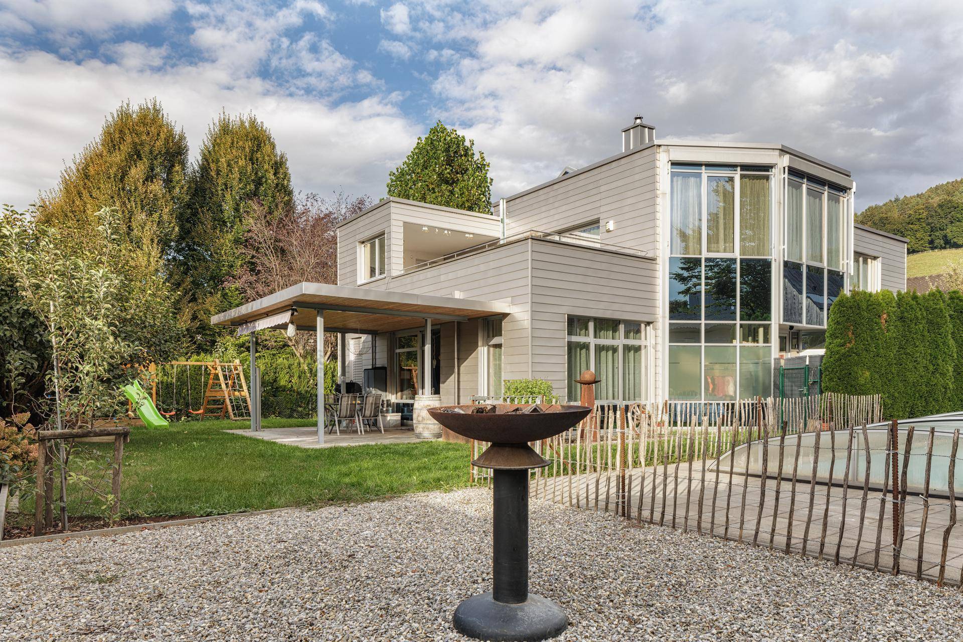 Großer Garten mit überdachter Terrasse, Spielplatz und Pool, umgeben von viel Grün und Bäumen.