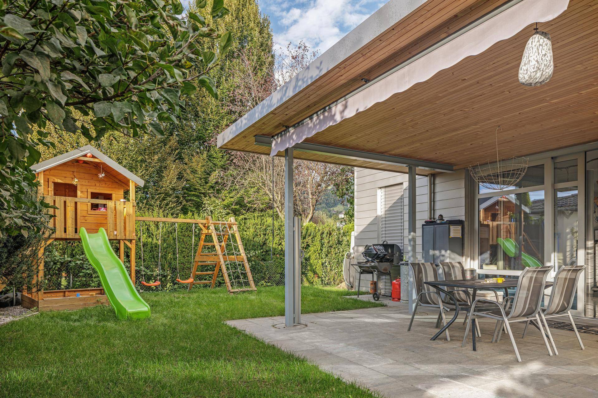Überdachte Terrasse mit Essbereich und angrenzendem Spielplatz im grünen Gartenbereich des Hauses.