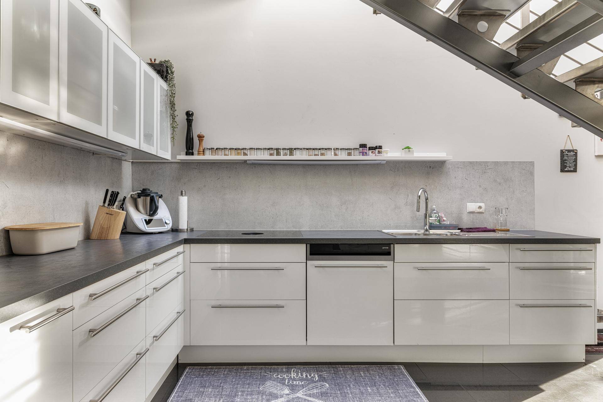 Moderne Küche mit weißen Hochglanzfronten, dunkler Arbeitsplatte und integrierten Geräten unter der Treppe.