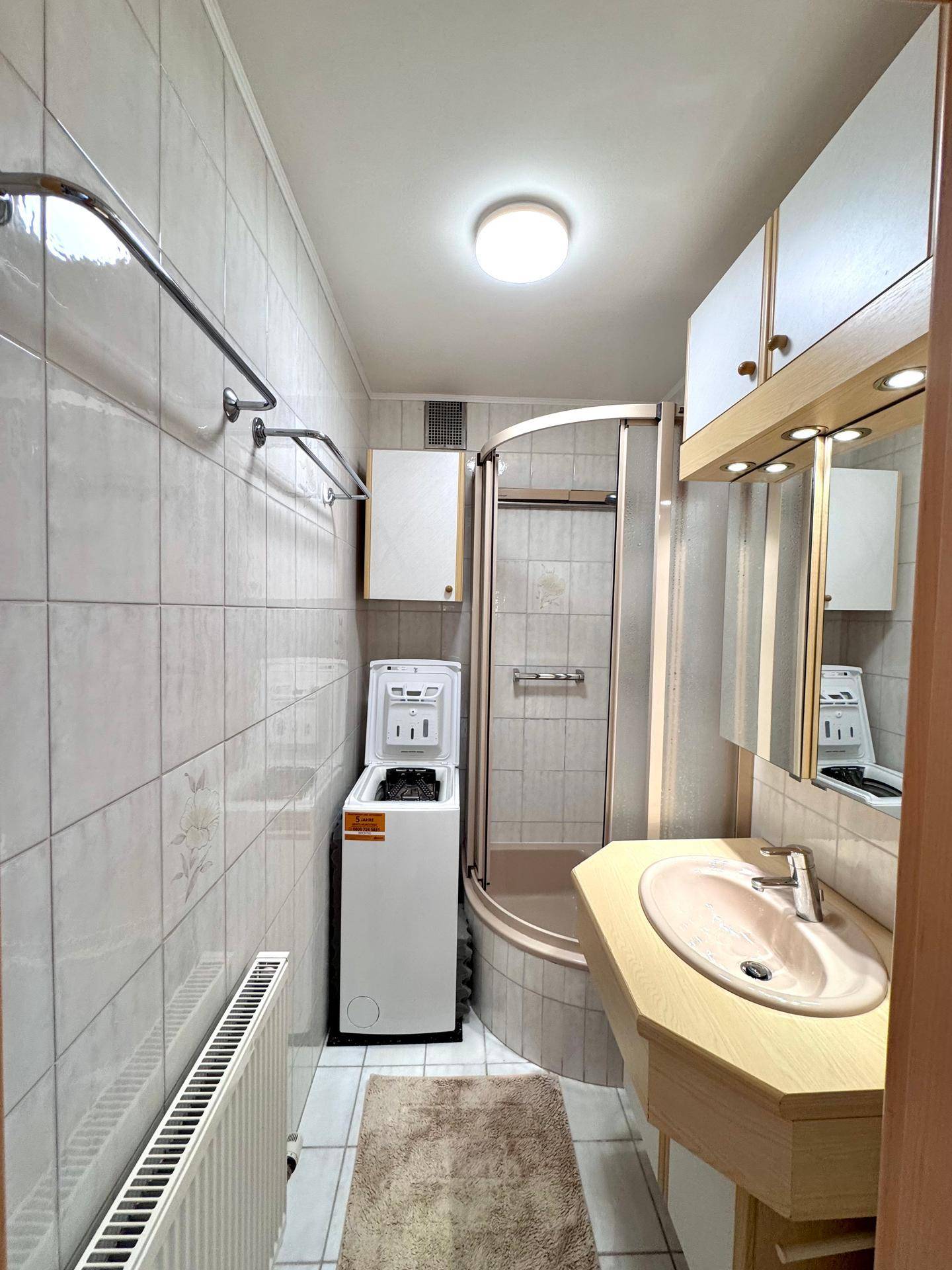 Gefliestes Badezimmer mit Duschkabine, Waschbecken und Waschmaschine.