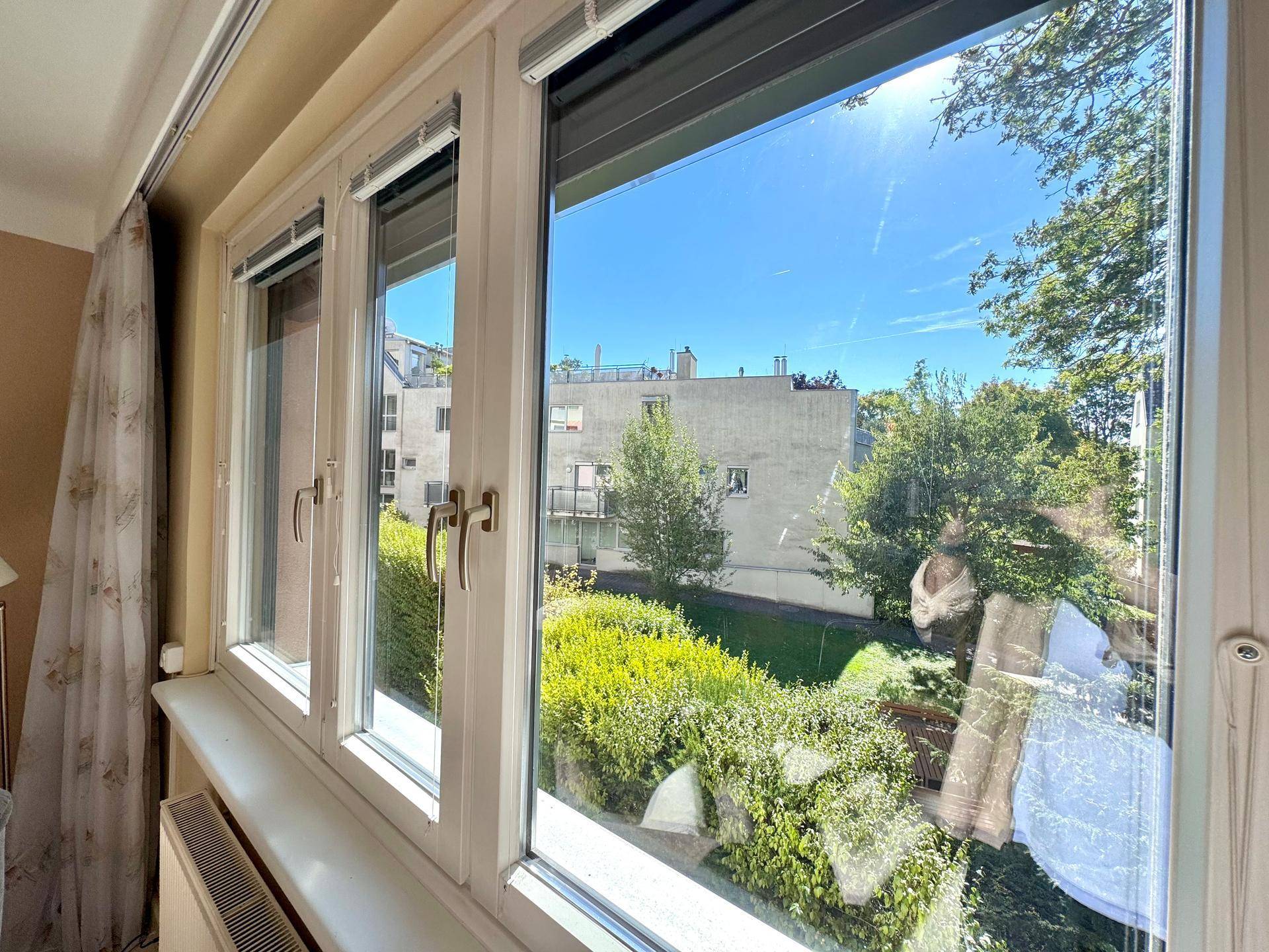 Fensterblick aus dem Wohnzimmer auf grüne Büsche und ein angrenzendes Gebäude.