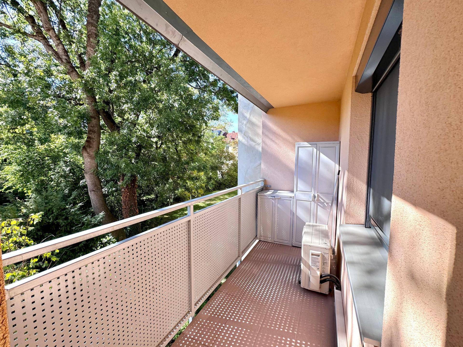 Heller Balkon mit Metallgeländer, Stauraum und Blick auf alte Bäume.