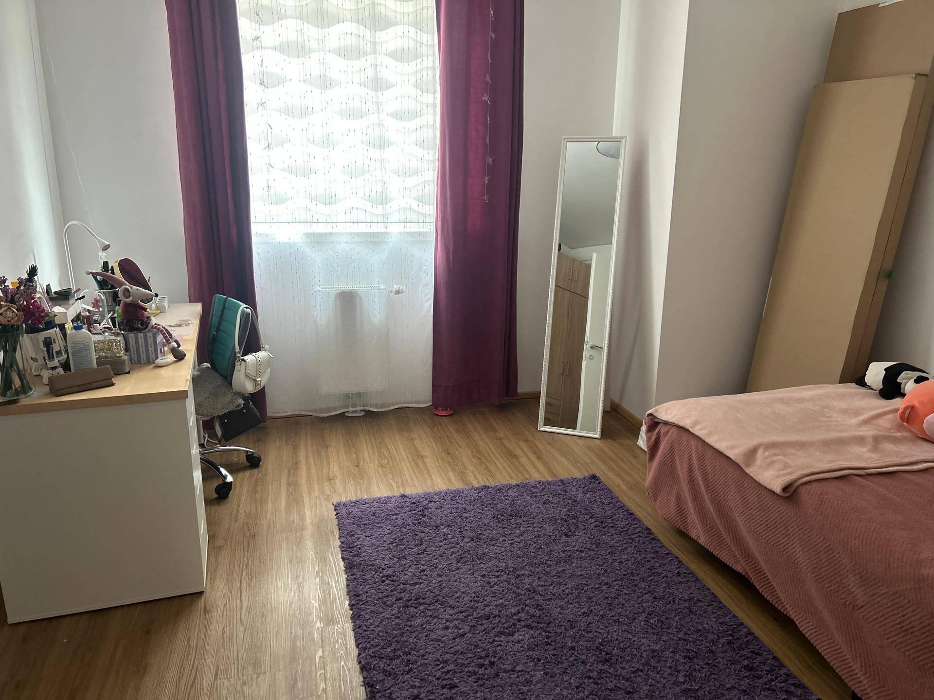 Helles Schlafzimmer mit Laminatboden, Schreibtisch, Bett und einem großen Fenster mit roten Vorhängen.