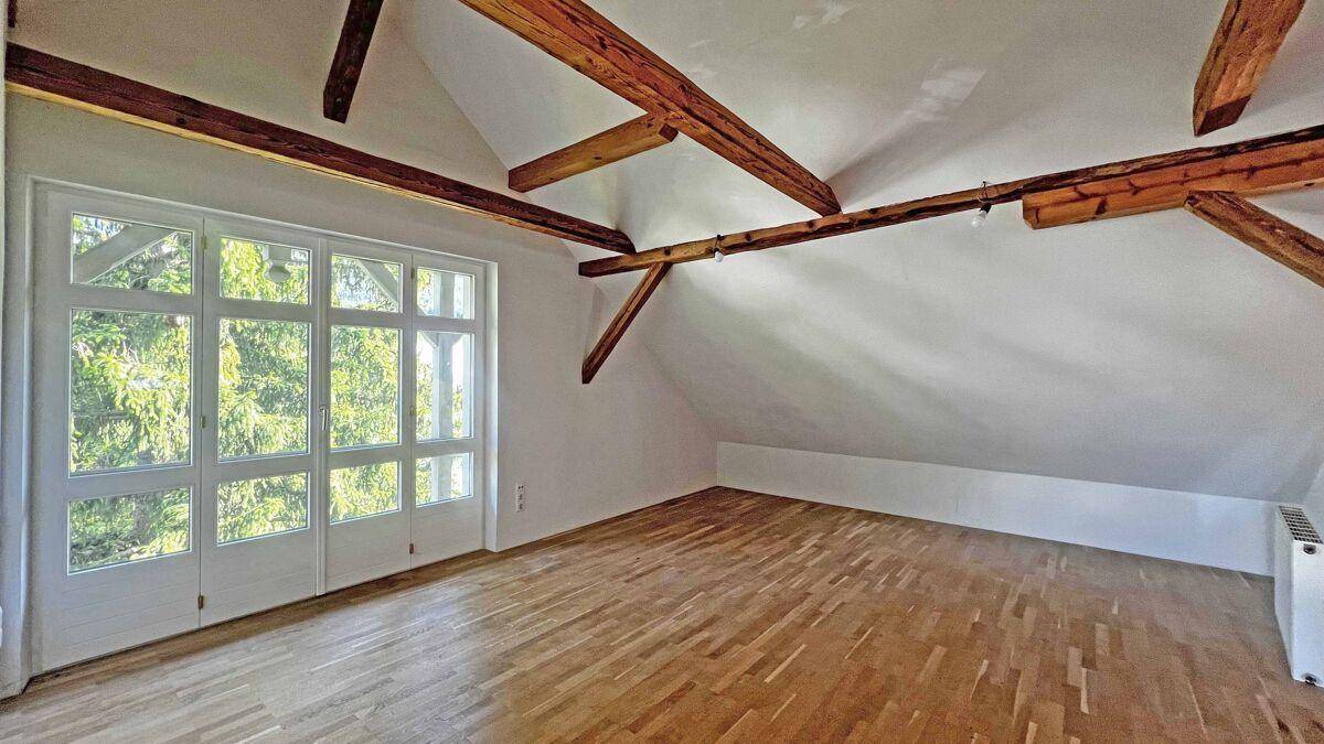 Geräumiges Dachzimmer mit Holzbalken, Parkettboden und großer Fensterfront.