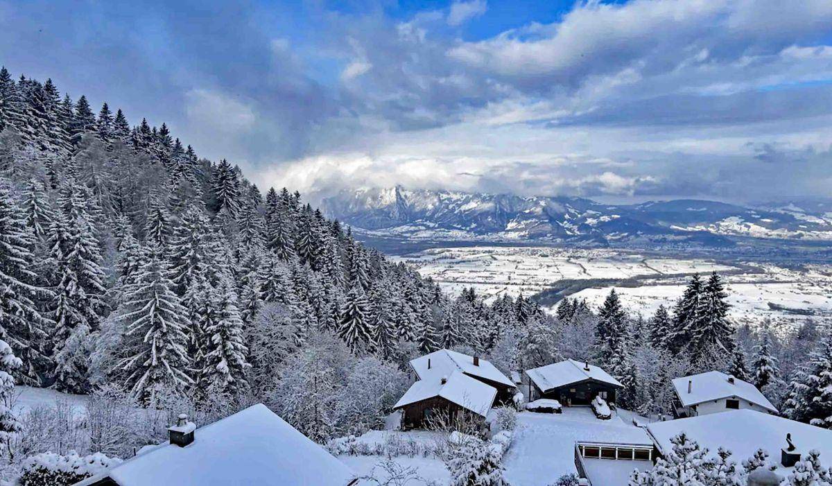 Winterlandschaft mit verschneiten Häusern und Bäumen, umgeben von einem weiten Tal und Bergen.