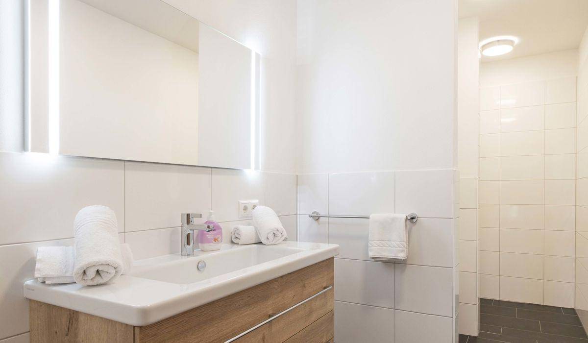 Modernes Badezimmer mit hellem Holzwaschtisch, beleuchtetem Spiegel und begehbarer Dusche.