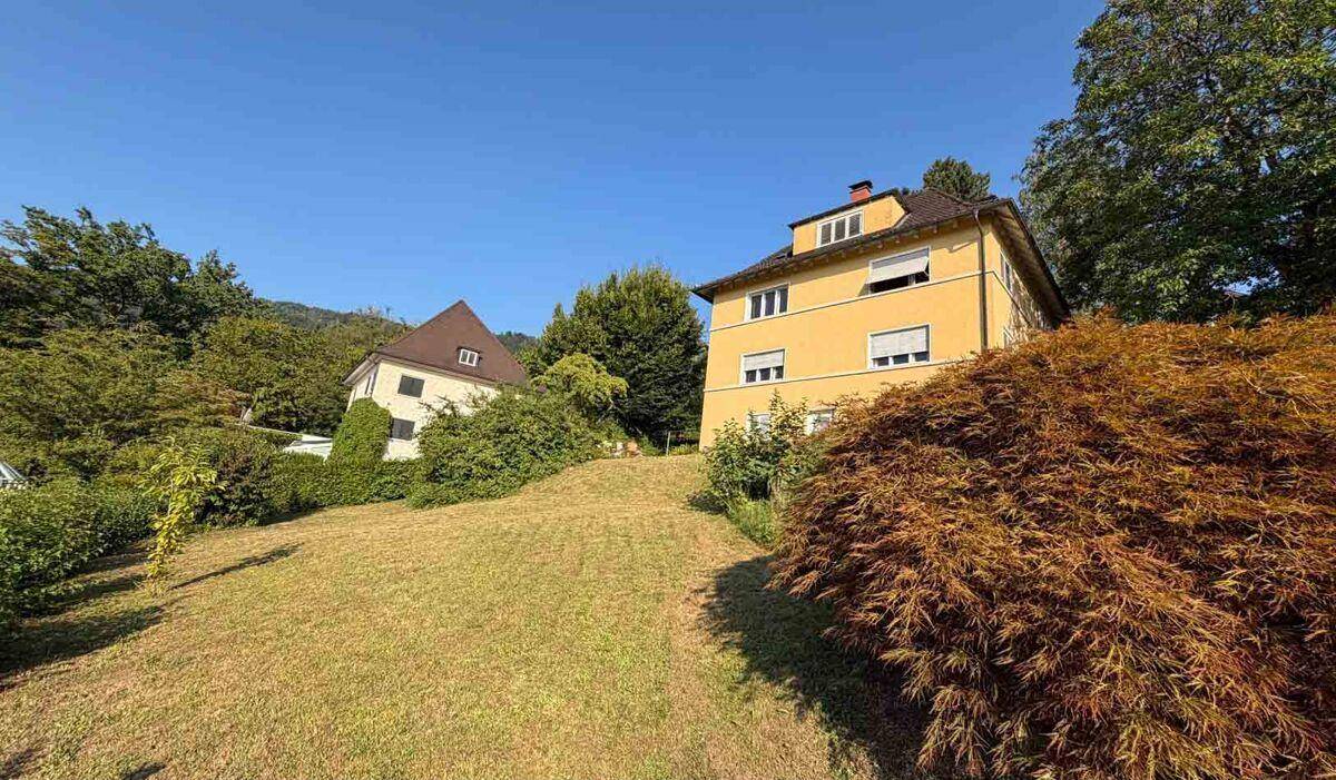 Gelbes Haus auf einem grünen Hügel mit weiteren Gebäuden und Bäumen im Hintergrund unter blauem Himmel.
