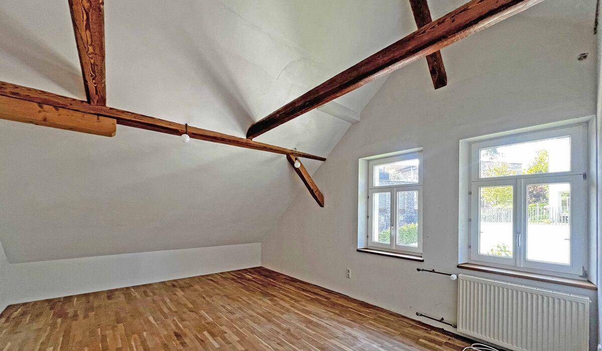 Helles Dachzimmer mit Holzbalken, Parkettboden und zwei Fenstern.