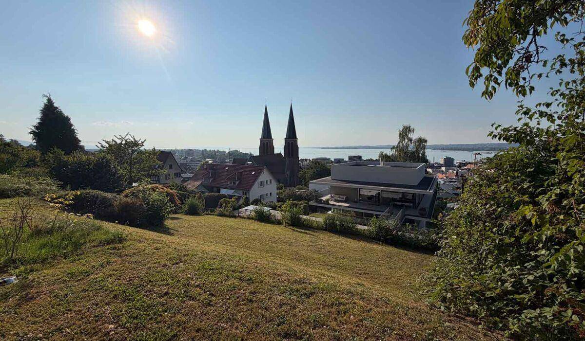 Panoramablick von einem grünen Hügel über eine Stadt mit Kirche und einem weiten See im Hintergrund.