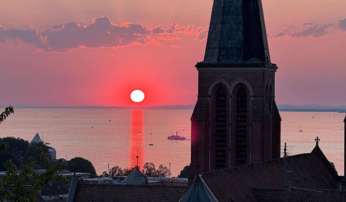 Spektakulärer Sonnenuntergang über einem See mit einem historischen Kirchturm im Vordergrund und rotem Himmel.
