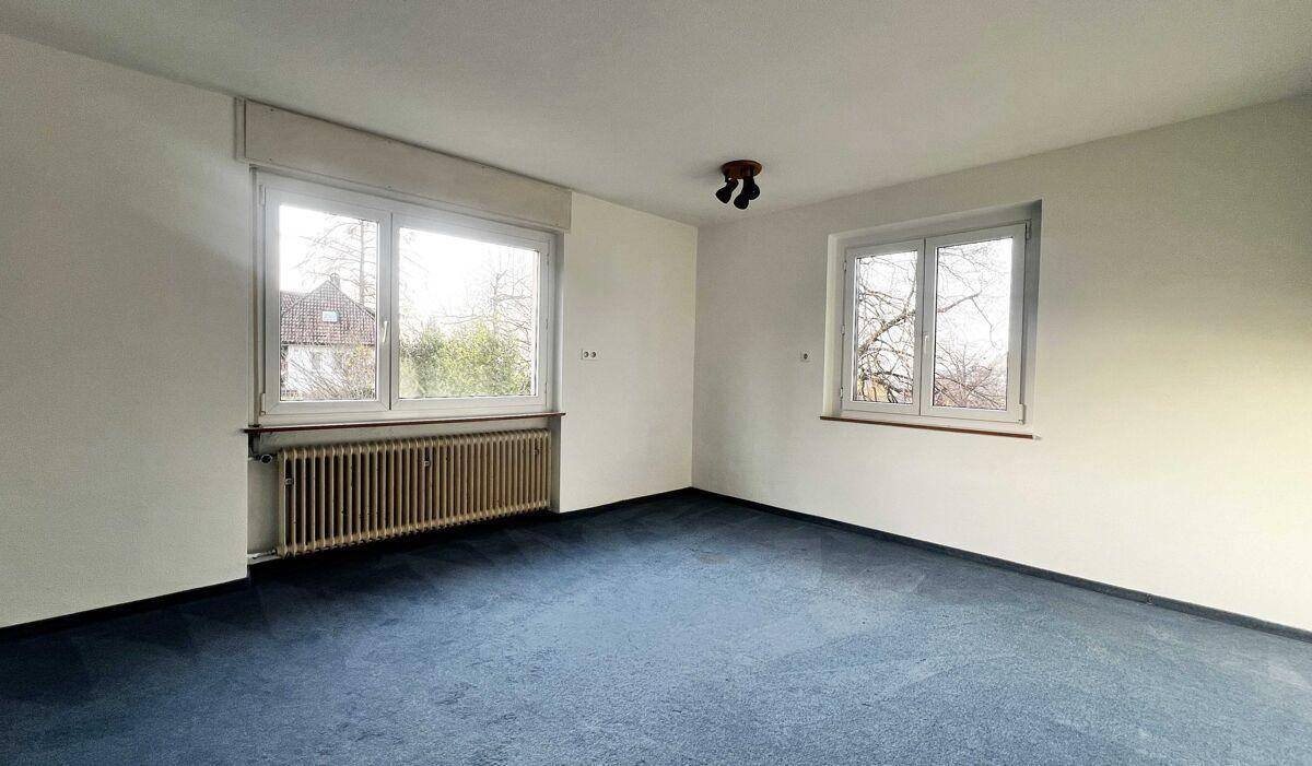 Schlafzimmer mit blauem Teppichboden und zwei Fenstern, die viel Tageslicht hereinlassen.