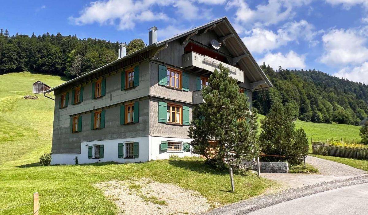 Traditionelles Alpenhaus mit grünen Fensterläden, Balkonen und einer Zufahrt, eingebettet in grüne Hügel.
