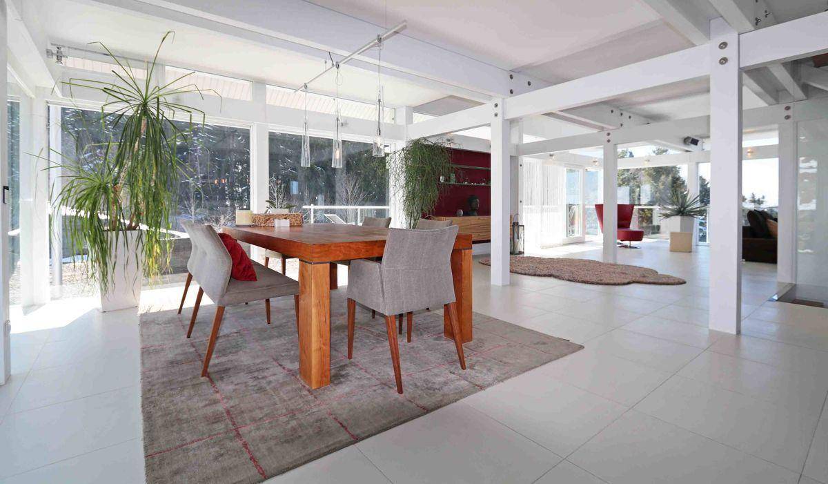 Heller Essbereich mit Holztisch und Stühlen, umgeben von großen Fenstern und Pflanzen.