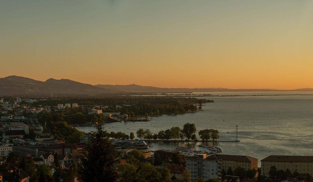 Panoramablick auf den See, die Stadt und die umliegenden Berge bei warmem Abendlicht.