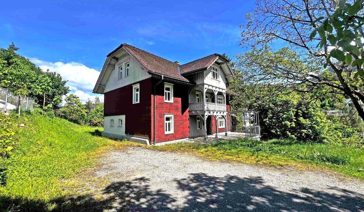 Traditionelles Haus mit roter Fassade und Holzbalkonen, umgeben von grüner Natur unter blauem Himmel.