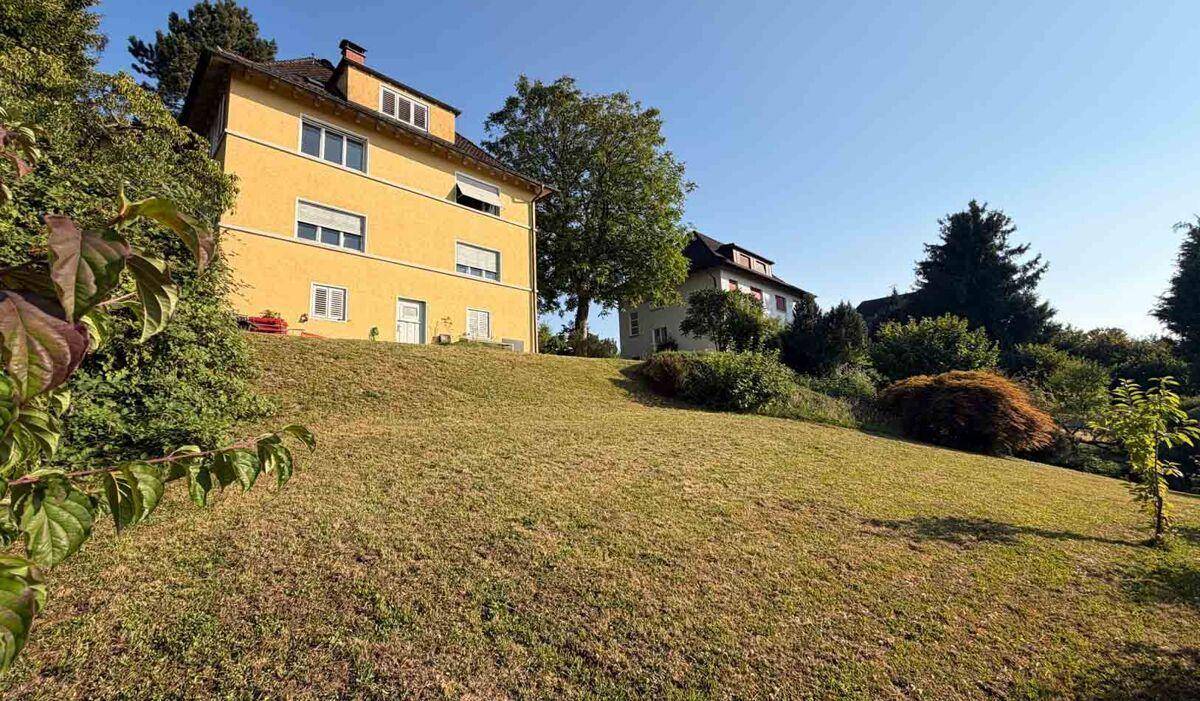 Gelbes Haus auf einem grünen Hang mit üppiger Vegetation und einem weiteren Gebäude im Hintergrund.