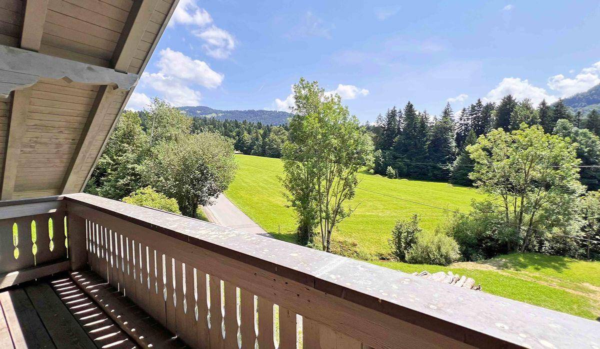 Holzbalkon mit weitem Blick über grüne Wiesen und bewaldete Hügel unter blauem Himmel.