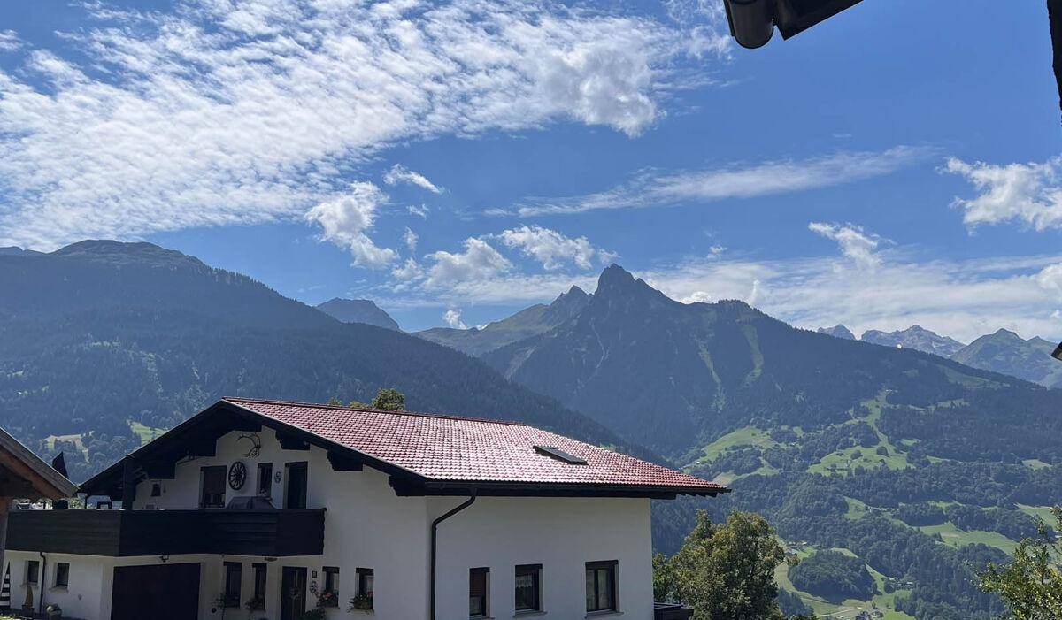 Weißes Haus mit rotem Dach und Balkon vor einer beeindruckenden Bergkulisse.