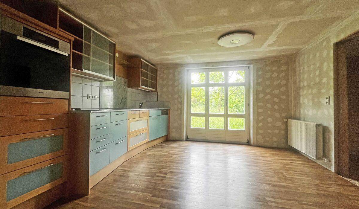 Küche mit Einbauschränken, integriertem Ofen und Holzboden. Die Wände sind im Rohzustand, mit großem Fenster zum Garten.