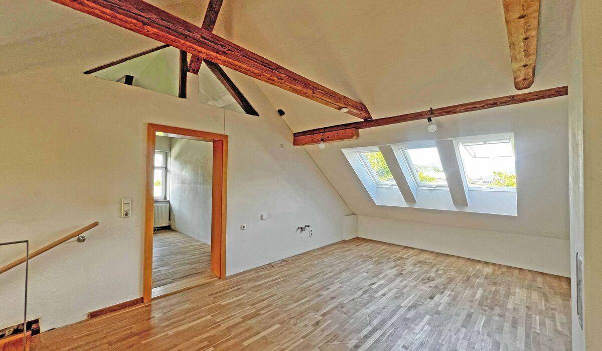 Dachzimmer mit schrägen Wänden, Holzbalken, Parkettboden und Dachfenstern.