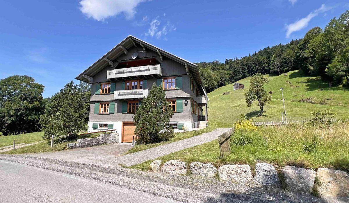 Traditionelles Alpenhaus mit grünen Fensterläden, Balkonen und einer Auffahrt, umgeben von Natur.