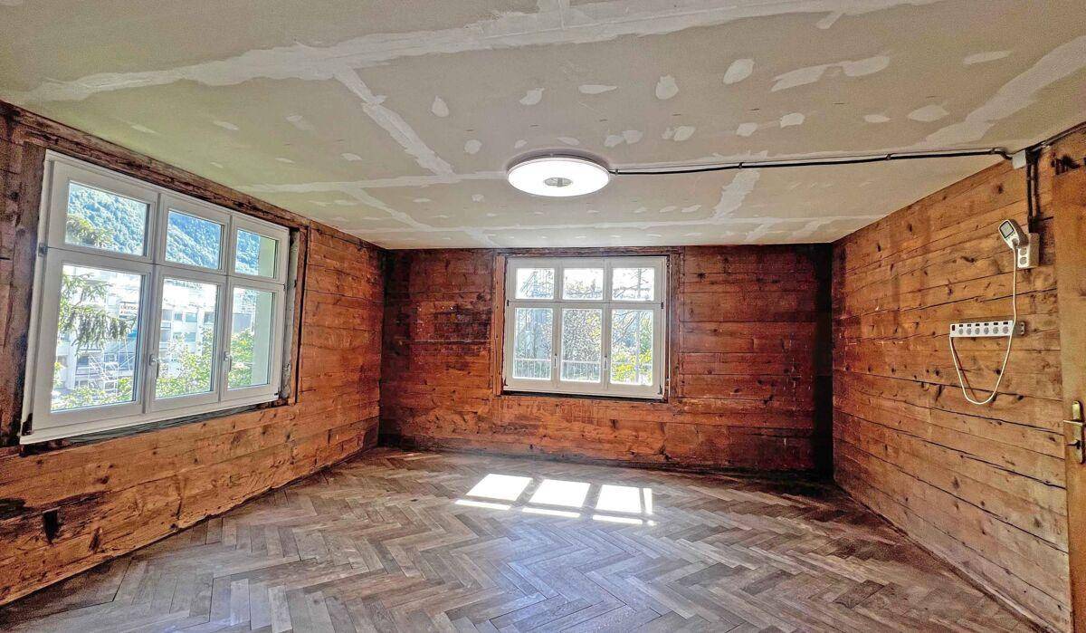 Helles Zimmer mit Holzwänden, Fischgrätparkett und zwei großen Fenstern.
