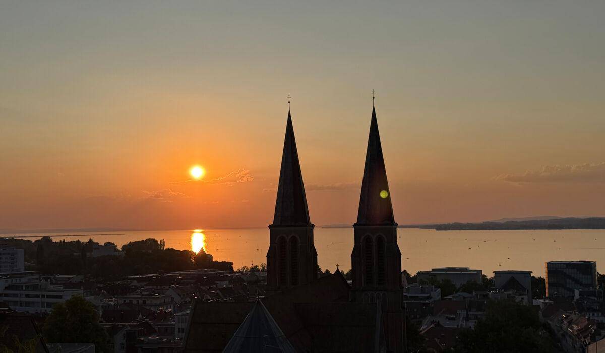 Malerischer Sonnenuntergang über einem See, eingerahmt von den Silhouetten zweier Kirchtürme.