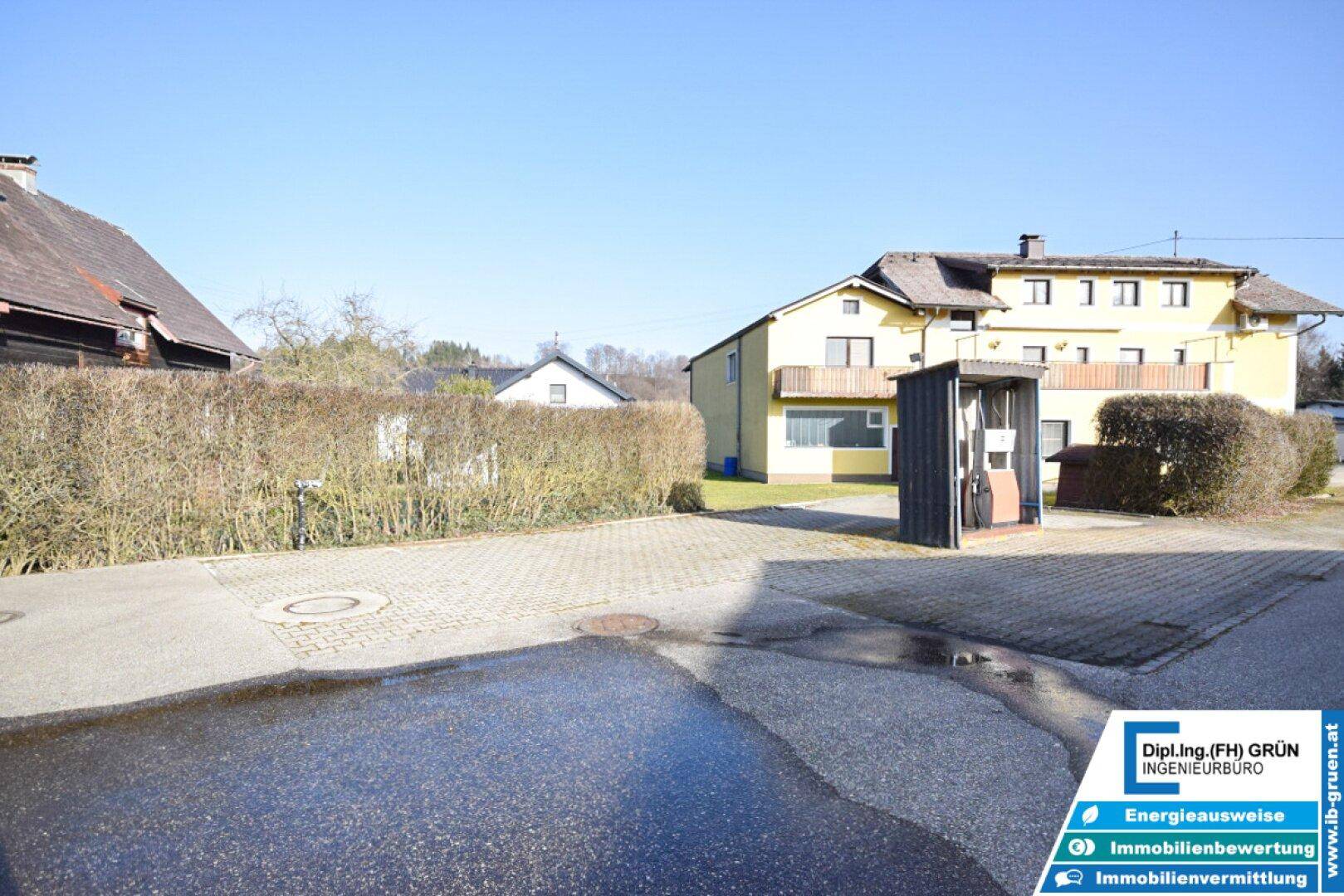 Außenbereich mit Hecke, alter Zapfsäule und Blick auf das gelbe Gebäude im Hintergrund.