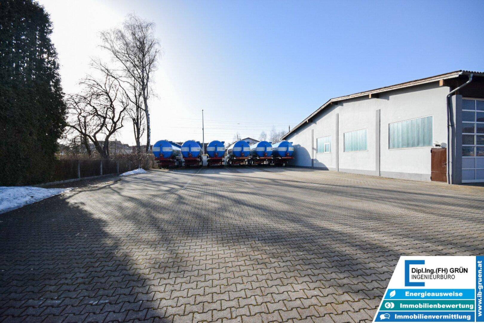 Gepflasterter Vorplatz vor der Halle mit abgestellten Tankwagen und Bäumen im Hintergrund.