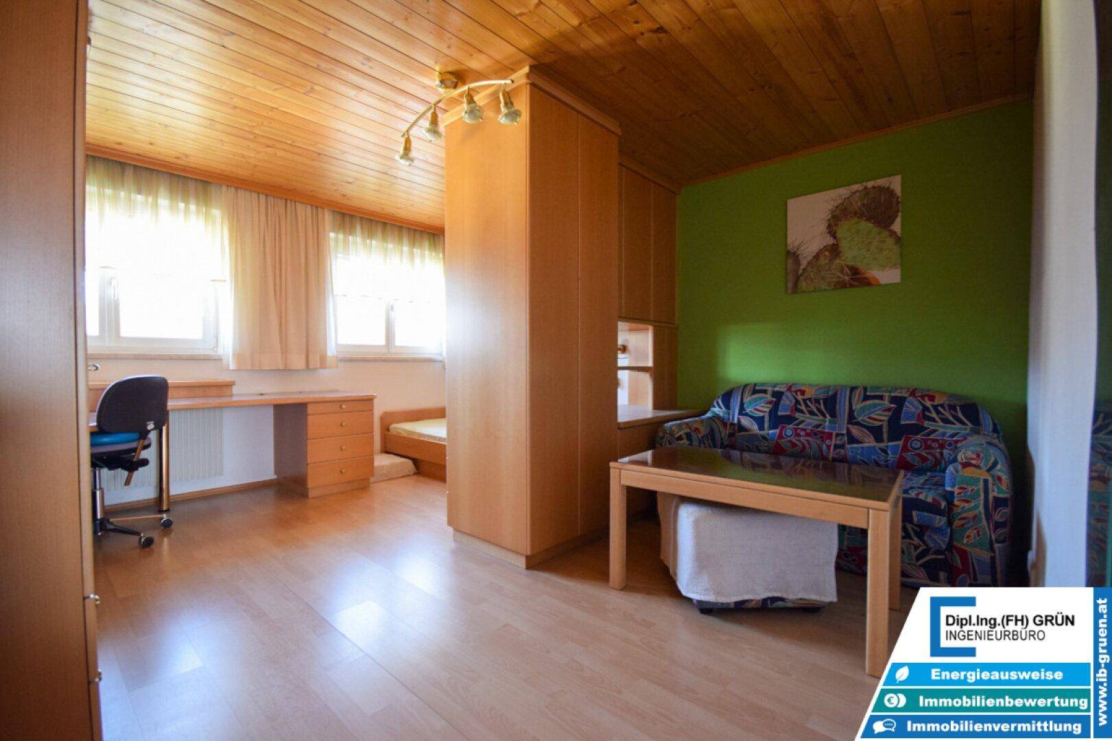 Helles Schlafzimmer mit Schreibtisch, Sofa und Holzdecke unter Dachschräge.