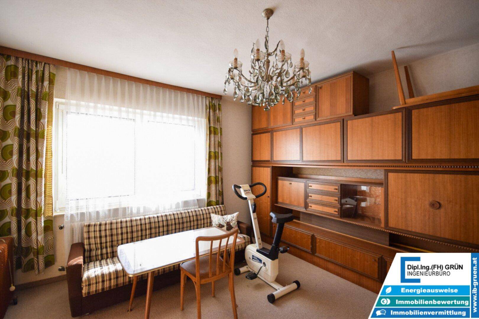 Wohnzimmer mit Sofa, Wandregal und Heimtrainer, beleuchtet von einem Kronleuchter.