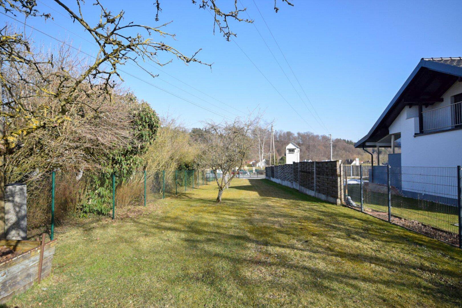 Weitläufiger Gartenbereich mit Rasenfläche, Zaun und Bäumen unter blauem Himmel.