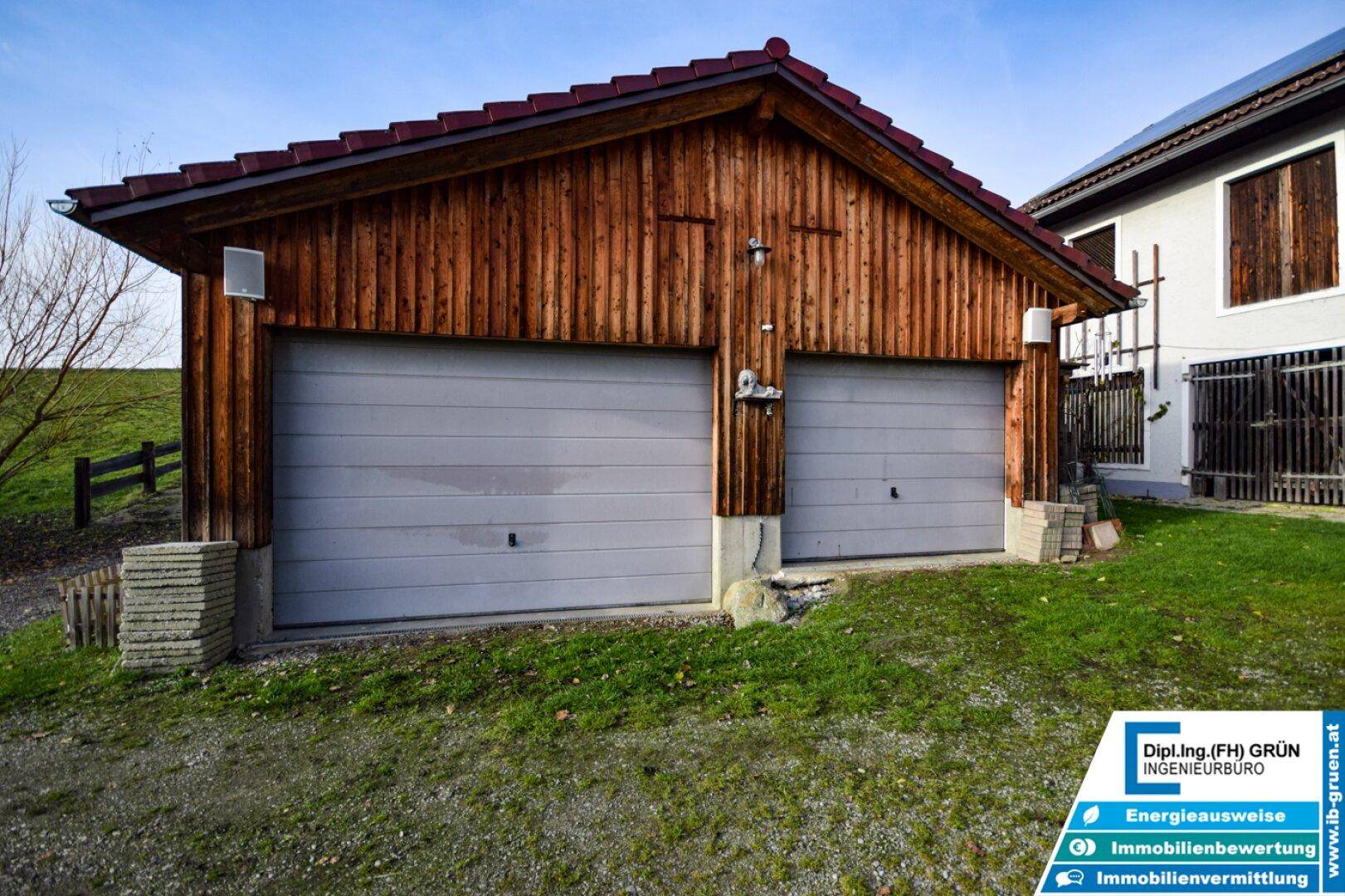 Doppelgarage aus Holz mit zwei grauen Toren und einem roten Ziegeldach, umgeben von Grün.